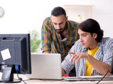 Two men are working together at a desk in an office. One man is seated and typing on a laptop, ensuring the software is up-to-date, while the other man is standing and looking at the laptop screen. Both are wearing casual clothes, and there are two computer monitors on the desk.