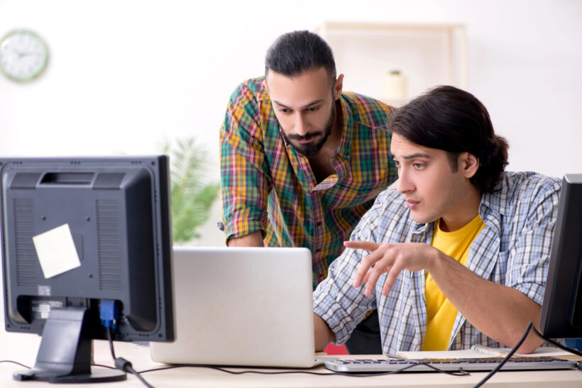 Two men are working together at a desk in an office. One man is seated and typing on a laptop, ensuring the software is up-to-date, while the other man is standing and looking at the laptop screen. Both are wearing casual clothes, and there are two computer monitors on the desk.