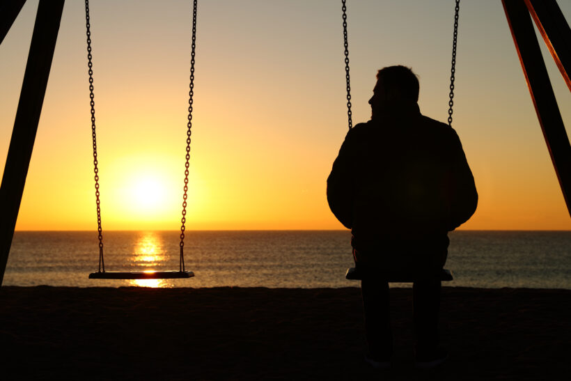 A person sits alone on a swing facing the ocean during sunset. The sun is low on the horizon, casting a warm glow over the water. Another swing, like a silent echo of their thoughts, is empty next to them. The scene has a serene and contemplative atmosphere, reminiscent of moments in "how i met my big brother.