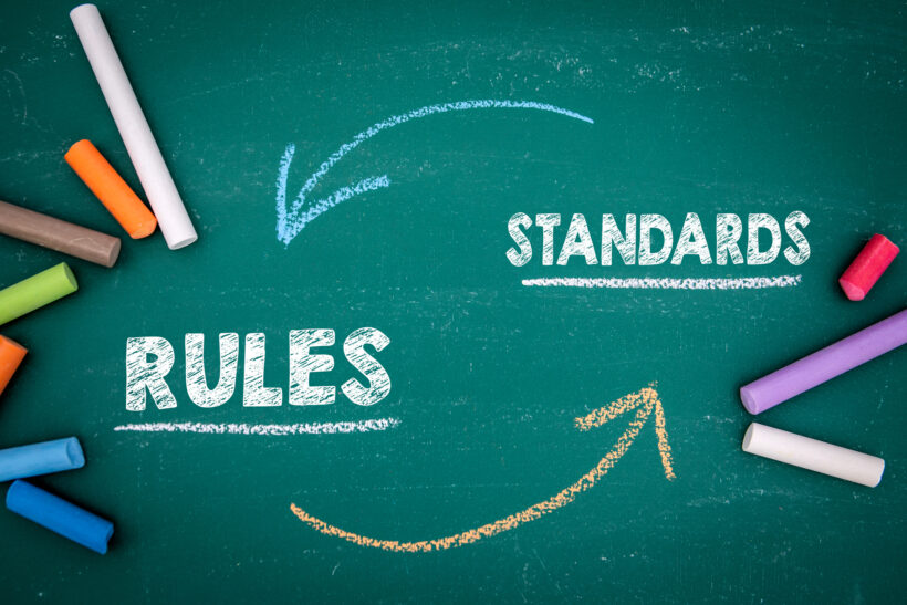 A green chalkboard with the words "rules" and "standards" written in white chalk, connected by arrows drawn in blue and orange chalk, illustrates the rigorous guidelines for pci-compliant web hosting. Several pieces of colored chalk are scattered around the board.