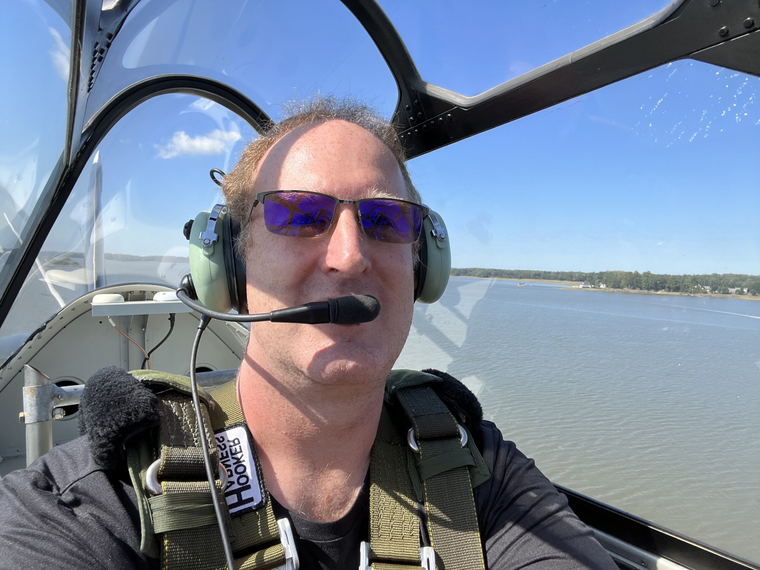 A person wearing aviator sunglasses, a headset with a microphone, and a green harness is seated in the cockpit of an aircraft. The photo is taken mid-flight with a body of water and distant shoreline visible in the background.