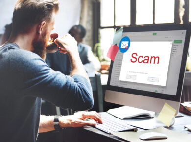 How To Recognize And Avoid Phishing Scams 3 A man is sitting at a desk in front of a computer. The screen displays a warning message with the word "scam" in large red letters, indicating potential phishing scams. The man is drinking from a cup while looking at the screen. The desk has various office supplies on it.