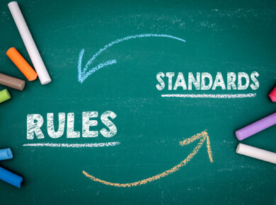 Do You Have Pci-Compliant Web Hosting? 5 A green chalkboard with the words "rules" and "standards" written in white chalk, connected by arrows drawn in blue and orange chalk, illustrates the rigorous guidelines for pci-compliant web hosting. Several pieces of colored chalk are scattered around the board.